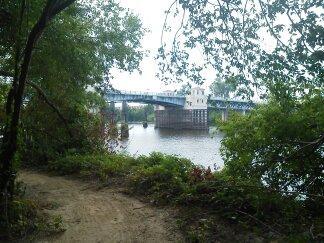 A scenic view of a riverbank framed by greenery, featuring a steel bridge crossing over the water in the background. The path leading towards the river is sandy, surrounded by lush plants and trees. Forsythe Park mountain bike trail.