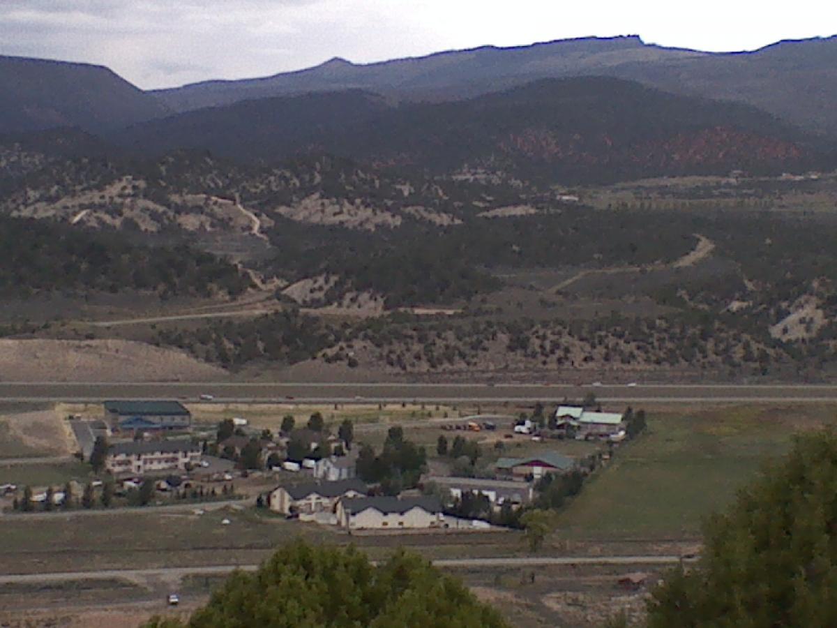 A scenic view of a rural landscape featuring a small town nestled in a valley, surrounded by rolling hills and mountains in the background. The image displays various buildings, including residential houses and commercial structures, with roads winding through the area. The sky is overcast, suggesting a cloudy day. The Boneyard mountain bike trail.