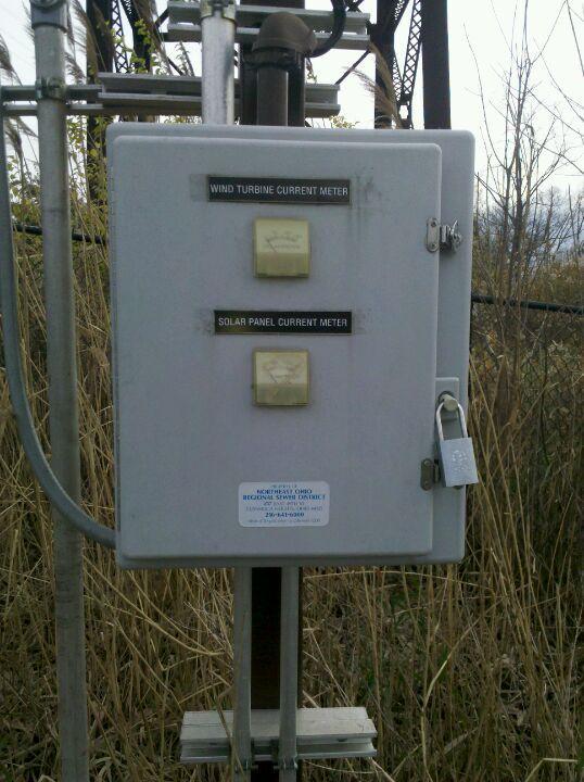 A metal electrical box mounted on a pole, featuring two labeled buttons for "Wind Turbine Current Meter" and "Solar Panel Current Meter." The box is secured with a padlock and displays a blue informational sticker at the bottom. The surrounding area includes tall grasses and a background of structures. Ohio And Erie Canal mountain bike trail.