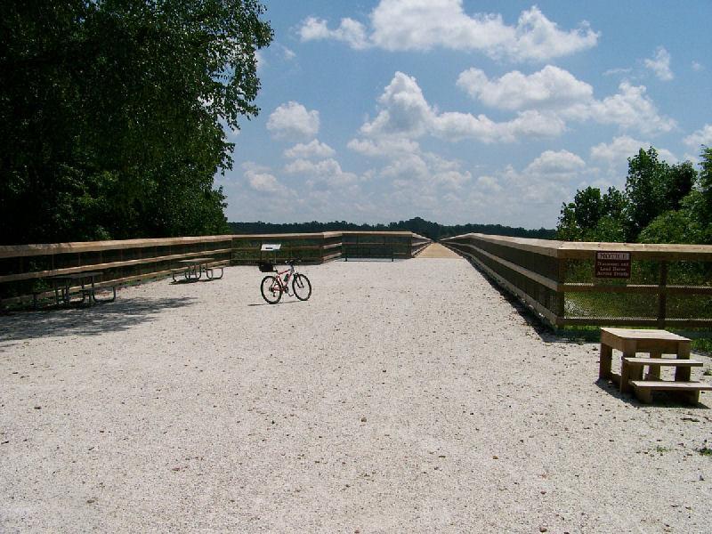 A wide, gravel pathway stretching into the distance, flanked by wooden railings and surrounded by lush greenery. A small pink bicycle is parked to the side, and a few benches and informational signs are visible along the route. The sky is bright with fluffy white clouds, creating a picturesque outdoor scene. High Bridge Trail mountain bike trail.