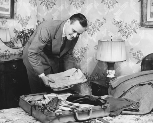A man in a suit smiles as he neatly folds and packs clothing into an open suitcase on a bed, surrounded by floral wallpaper and a lamp.