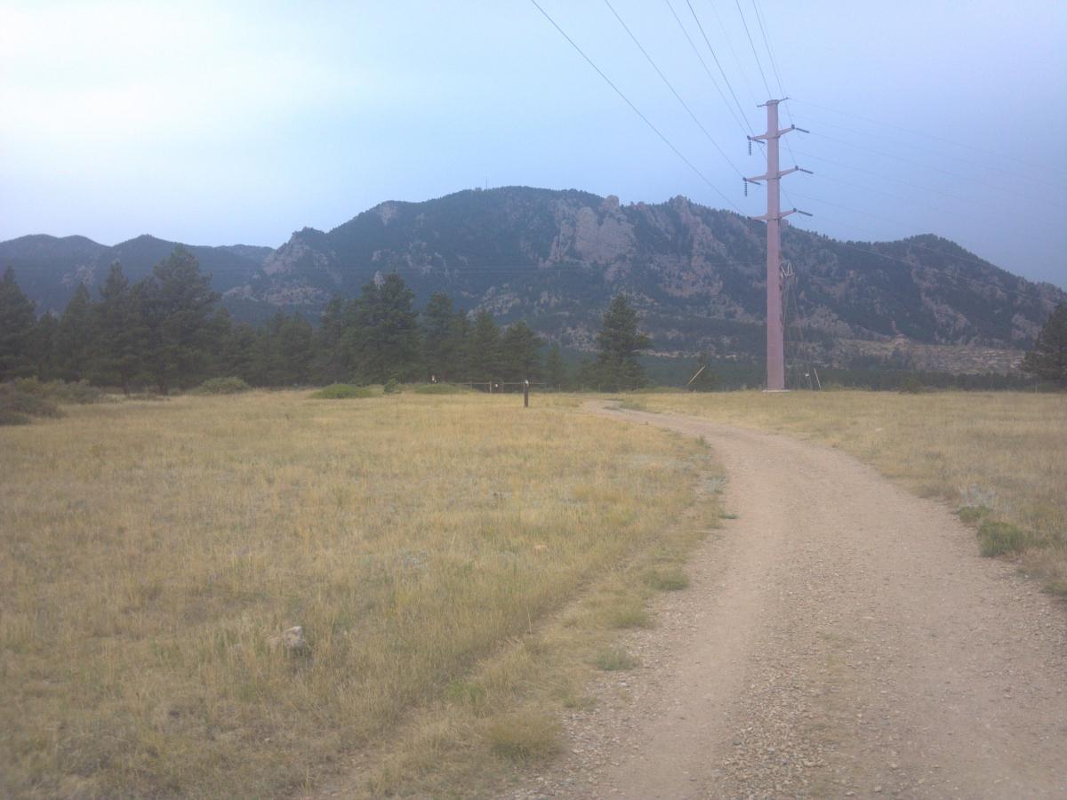 A dirt path winds through a grassy field, leading towards a mountainous backdrop under a cloudy sky. Tall pine trees are scattered throughout the area, with a prominent pink power line tower standing alongside the path. The mountains in the distance show rugged terrain and are partially obscured by clouds. Flatirons Vista mountain bike trail.