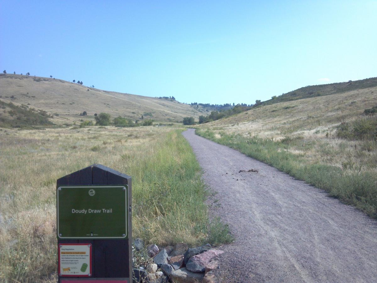 A scenic view of the Doudy Draw Trail, featuring a dirt path that winds through an open grassy landscape, bordered by rolling hills under a clear blue sky. In the foreground, a trailhead sign indicates the trail name and provides information for hikers. Doudy Draw Trail mountain bike trail.
