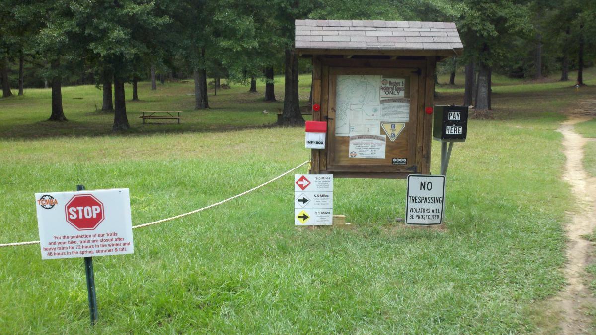 Signage and information kiosk at a park entrance, surrounded by grassy areas and trees. The kiosk features a trail map with designated distances, and signs include warnings about trail closures after heavy rain, a stop sign, and a pay station for entry. A "No Trespassing" sign indicates that violators will be prosecuted. A picnic table is visible in the background. The Ridgeland Trails mountain bike trail.
