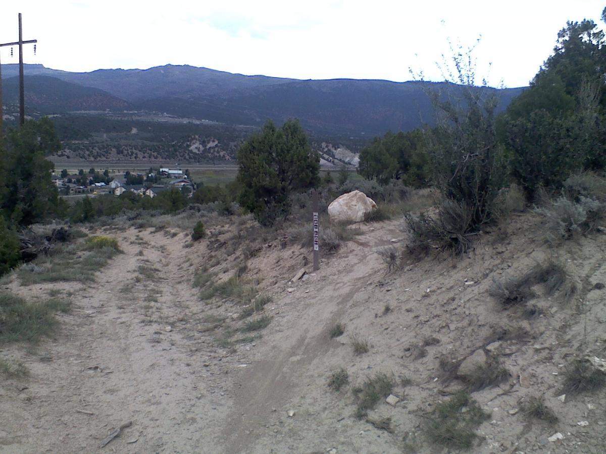 An unpaved trail winds through a landscape of sparse vegetation and dirt, leading down a slope towards a small town in the valley below. In the foreground, a wooden sign marks the trail, and a large rock is positioned near the path. In the background, rolling mountains rise under a cloudy sky. Electrical poles are visible on the left side of the image. The Boneyard mountain bike trail.