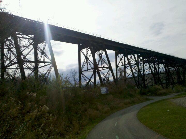 Alt text: A tall, industrial-style railway bridge rises above a path surrounded by greenery, with a bright sun emitting light from behind the structure. The sky is partly cloudy, and the foreground features a winding walkway. Ohio And Erie Canal mountain bike trail.