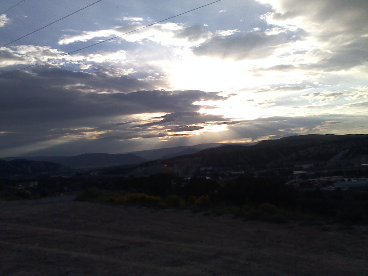 A scenic view of a mountainous landscape under a dramatic sky, featuring a mix of clouds and rays of sunlight breaking through. The sun is located behind the clouds, casting a gentle glow over the hills and valleys below. The foreground shows a gravelly area, with hints of vegetation, while the distant mountains silhouette against the soft light, creating a tranquil atmosphere. The Boneyard mountain bike trail.