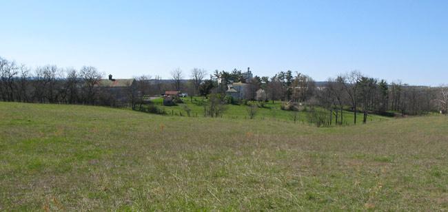 A wide-open field with green grass leading to a distant view of several houses nestled among trees under a clear blue sky. 3rd Battle Of Winchester Trail mountain bike trail.