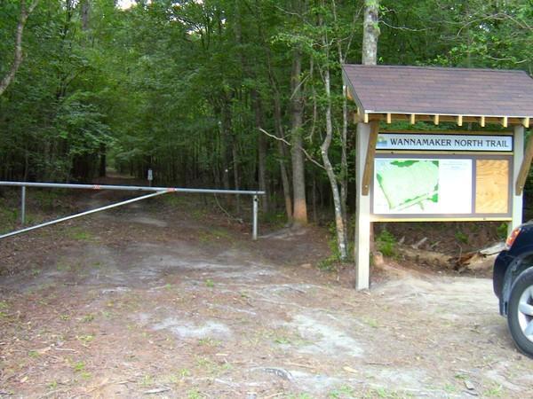 A quiet dirt path leading into a wooded area, marked by a wooden sign indicating the "Wannamaker North Trail." The sign displays a map of the trail and is next to a vehicle parked on the side. There is a barrier across the trail entrance, and lush green trees surround the entrance, creating a serene outdoor atmosphere. Wannamaker Park mountain bike trail.