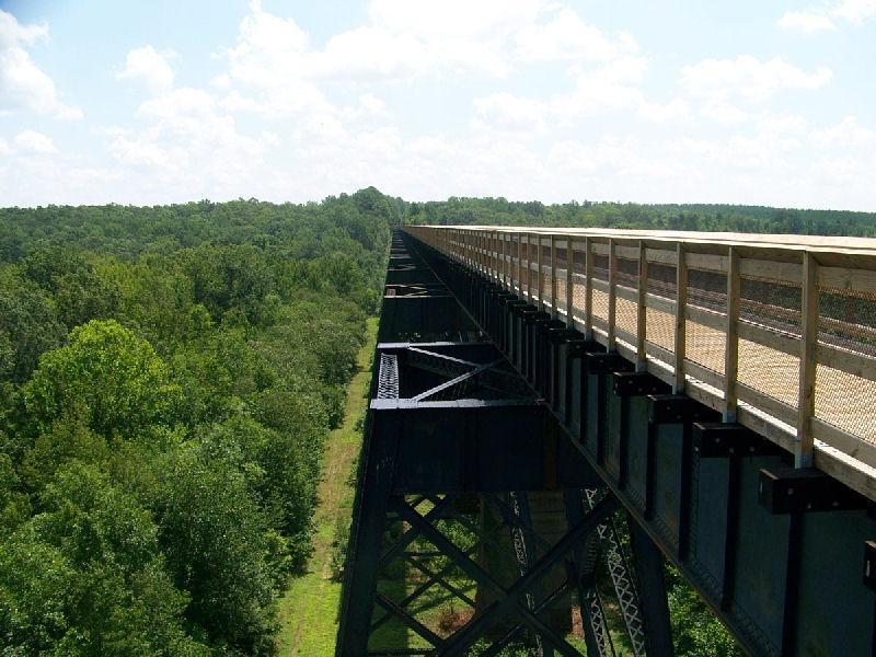 A high-angle view of a long, elevated bridge stretching over a dense forested area, with green trees visible below and a bright, partly cloudy sky above. The bridge features a wooden walkway bordered by safety railings. High Bridge Trail mountain bike trail.