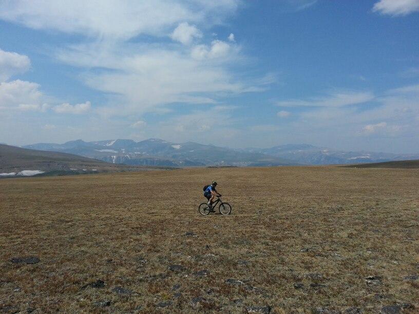 A cyclist riding on a vast, dry grassy landscape with distant mountains under a blue sky scattered with clouds. Line Creek Plateau mountain bike trail.