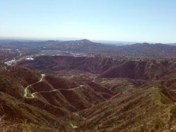 A panoramic view of rolling hills and valleys under a clear blue sky, with winding roads visible throughout the landscape. The scene captures the natural beauty of the terrain, showcasing a mix of greenery and dry elements typical of hilly regions. El Prieto mountain bike trail.