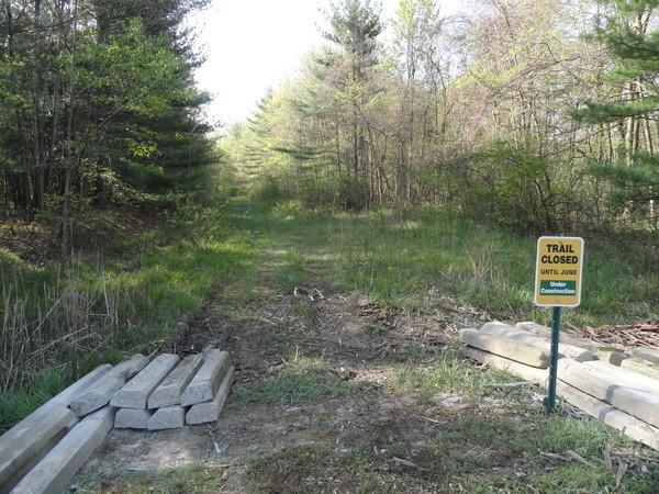 A narrow, grassy path through a wooded area is blocked by large wooden logs and a sign stating "Trail Closed Until June." Lush green trees surround the trail, indicating a natural, outdoor environment. Royalview mountain bike trail.