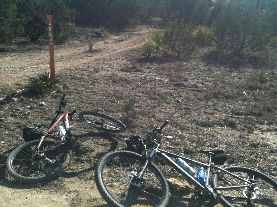Two mountain bikes lying on the ground next to a trail marker in a natural landscape. The terrain is dry with sparse vegetation and rocky patches. The bikes are positioned at an angle, showing their wheels and gear. The trail marker, painted orange, indicates the trail