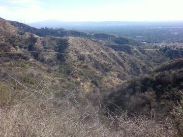 A panoramic view of rolling hills and valleys under a clear sky, showcasing dry, brown vegetation and a winding road in the distance. The landscape stretches towards a distant mountainous horizon, with hints of a city visible below. El Prieto mountain bike trail.
