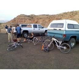 A group of people gathered near two parked vehicles, one is a silver pickup truck and the other is an older blue and white truck. Several mountain bikes are leaned against the ground, and some individuals are sitting on camping chairs while others are standing, chatting amidst a backdrop of rocky terrain. The scene captures a relaxed outdoor activity setting. Gunny Loop mountain bike trail.