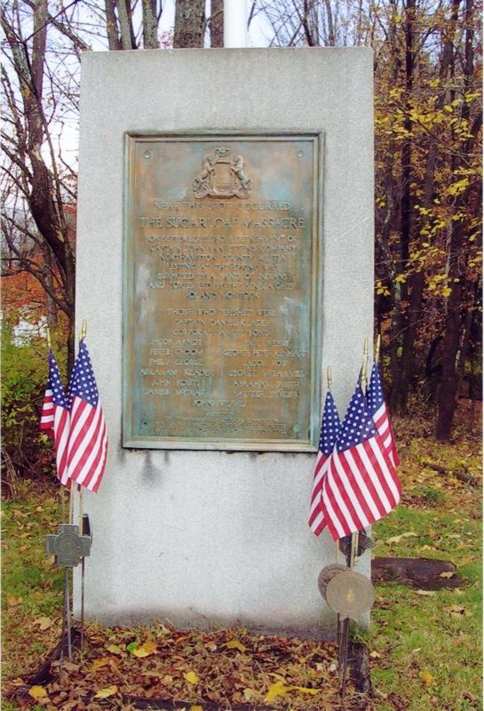 A granite monument commemorating the Sugarloaf Massacre, featuring a bronze plaque with historical inscriptions. The plaque is surrounded by small American flags and is set against a backdrop of trees with autumn foliage. Sugarloaf Mountain Bike Area mountain bike trail.