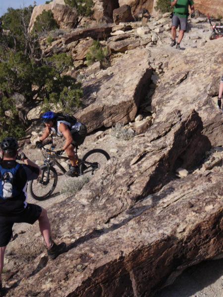 A mountain biker navigates rocky terrain, surrounded by other cyclists observing the trail. The scene showcases the rugged landscape and the challenge of off-road biking. Pet - A - Kes mountain bike trail.