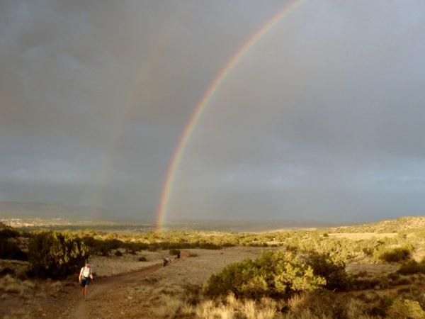 A scenic landscape featuring a dirt path winding through a grassy area with sparse shrubs. In the background, a dramatic sky showcases a double rainbow against a backdrop of dark, cloudy weather. Two hikers are seen walking along the path, with the vibrant rainbow creating a contrast to the subdued colors of the surrounding landscape. Gunny Loop mountain bike trail.