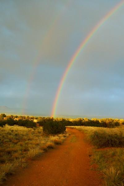 A vibrant double rainbow arcs over a dirt pathway surrounded by grassy fields and low shrubs, under a cloudy sky. The scene captures the beauty of nature after a rain shower. Gunny Loop mountain bike trail.