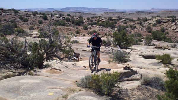 Mountain biker navigating rocky terrain in a desert landscape, surrounded by sparse vegetation and distant hills under a clear sky. Klondike Bluffs mountain bike trail.