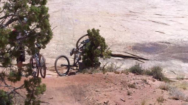A cyclist's mountain bike lying on its side next to a bush, with another bike and rocky terrain visible in the background. The scene captures a sunny outdoor setting, highlighting the natural landscape. Klondike Bluffs mountain bike trail.