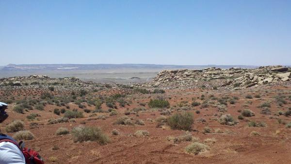 Wide panoramic view of a desert landscape featuring reddish-brown earth and sparse vegetation, with distant hills under a clear blue sky. Klondike Bluffs mountain bike trail.