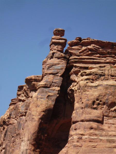 A rock formation characterized by a tall, slender column of stone topped with a smaller, round boulder. The formation is set against a clear blue sky, showcasing the textured layers of reddish-brown rock. Klondike Bluffs mountain bike trail.