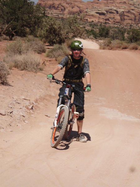 A mountain biker standing on a dirt trail in a rocky desert environment, wearing protective gear, a helmet, and casual cycling attire. The biker is smiling and holding onto their bike, with dry vegetation and rocky formations in the background. Klondike Bluffs mountain bike trail.