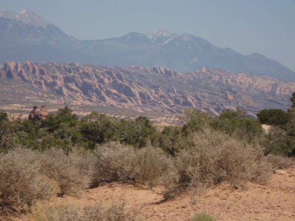 A panoramic view of rugged rock formations and rolling mountains under a clear sky, surrounded by sparse vegetation and desert landscape. The scene showcases the natural beauty of the terrain, highlighting layers of red rock and distant snow-capped peaks. Klondike Bluffs mountain bike trail.