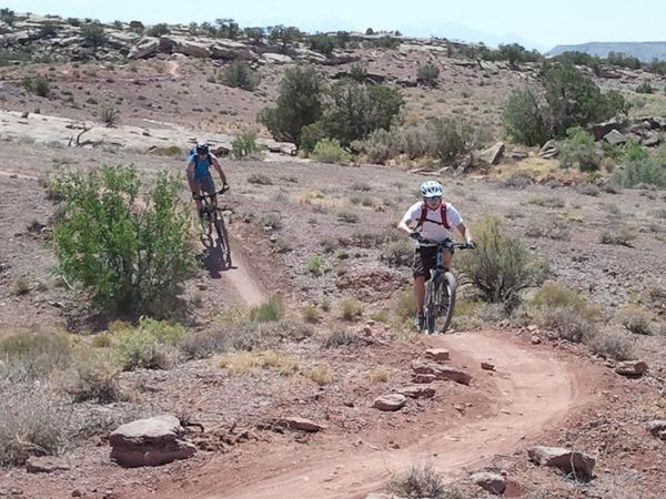 Two mountain bikers riding on a dirt trail in a rugged, arid landscape. The trail winds through rocky terrain with sparse vegetation and shrubs, under a clear blue sky. One biker is climbing the trail, while the other is positioned further up, adding a sense of motion and adventure to the scene. Little Salty mountain bike trail.