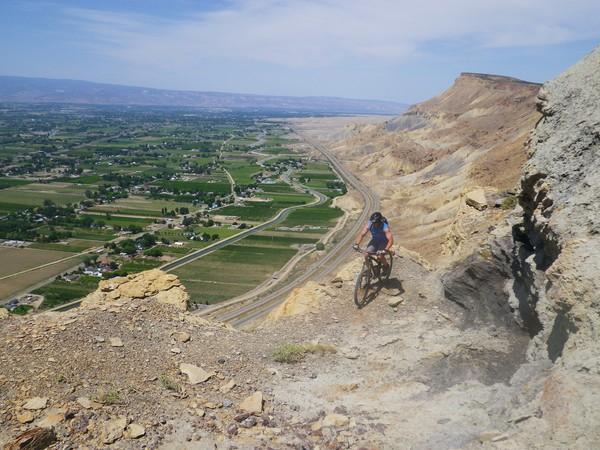 A mountain biker navigating a rocky trail on a steep cliff, overlooking a lush valley with fields and winding roads below. The sky is clear and blue, enhancing the scenic view of the landscape. Stagecoach mountain bike trail.