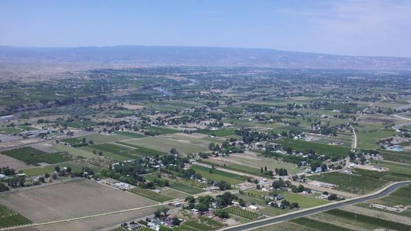 Aerial view of a rural landscape featuring farmlands, fields, and small communities. The area shows a patchwork of agricultural land with greenery, roads, and a distant mountain range under a clear blue sky. Stagecoach mountain bike trail.