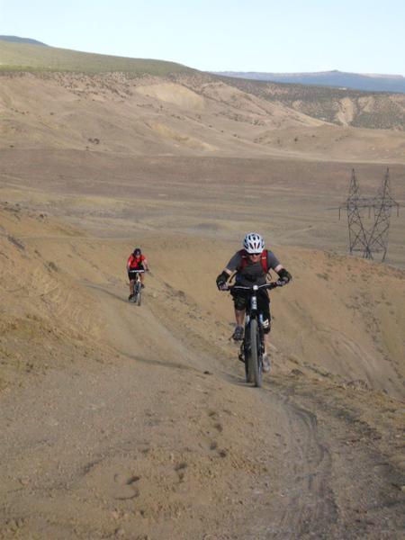 Two mountain bikers riding along a dirt trail on a hilly terrain, with a vast landscape of dry hills in the background and power lines visible in the distance. Horse Mountain mountain bike trail.