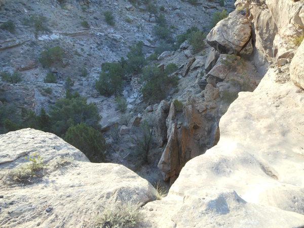A rocky cliff edge overlooking a steep, rugged canyon with sparse vegetation and scattered trees. The image captures the texture of the rocks and the natural landscape surrounding the gorge. Gunny Loop mountain bike trail.