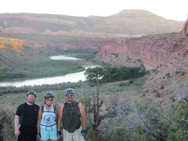 Three people stand together on a hillside overlooking a river and canyon. The landscape features red rock formations and green vegetation. The sun is setting, casting warm light on the scene. Two individuals are wearing helmets, suggesting they are biking, while everyone is dressed in casual outdoor clothing. Rustlers Loop mountain bike trail.