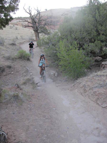 Two cyclists navigating a dusty trail in a natural outdoor setting. The first cyclist, wearing a helmet and casual attire, is riding downhill, while the second cyclist, dressed in black, is further back on the path. Surrounding vegetation includes shrubs and scattered trees, with rocky terrain visible along the trail. Rustlers Loop mountain bike trail.