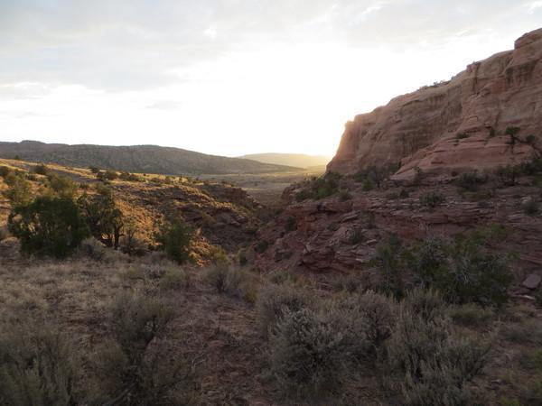 A scenic landscape featuring rocky cliffs and a valleys, illuminated by soft sunlight at sunset. The foreground includes shrubs and small trees, while the background showcases rolling hills and distant mountains under a partly cloudy sky. The warm glow of the setting sun enhances the natural colors of the terrain. Mary's Loop / Horsethief Bench mountain bike trail.
