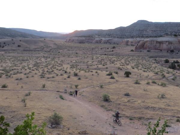 A scenic view of a vast, arid landscape during sunset, featuring rolling hills and rugged terrain. Two people can be seen walking along a dirt trail, while another person rides a bicycle. Sparse vegetation, including bushes and small plants, is scattered across the dry ground. The sky is painted with soft hues as the sun begins to set behind the mountains in the background. Mary's Loop / Horsethief Bench mountain bike trail.