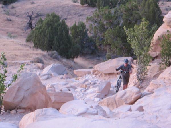 A mountain biker navigating a rocky trail surrounded by sparse vegetation and trees on a rugged terrain during dusk. The rider is pushing their bike over large stones and uneven ground. Mary's Loop / Horsethief Bench mountain bike trail.