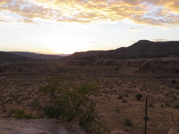 Sunset over a vast desert landscape, featuring rolling hills and scattered vegetation. The sky is filled with soft clouds illuminated by warm hues of orange and yellow, casting a serene glow across the terrain. Mary's Loop / Horsethief Bench mountain bike trail.