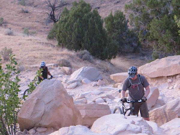 Two mountain bikers navigate a rocky terrain in a natural outdoor setting. One rider is seen pushing their bike over large stones, while the other is further up the trail, surrounded by shrubs and trees. The landscape is dry and hilly, indicating a rugged biking trail. Mary's Loop / Horsethief Bench mountain bike trail.