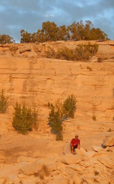 A person wearing a red shirt sits on rocky terrain at the base of a sandstone cliff, surrounded by sparse vegetation, under a partly cloudy sky. The warm tones of the rock formations are highlighted by the setting sun. Kokopelli Area Trails mountain bike trail.