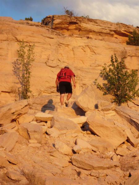 A person in a red shirt and black shorts is climbing a rocky, sandy hillside, with sparse vegetation and a backdrop of sandstone cliffs under a partly cloudy sky. Kokopelli Area Trails mountain bike trail.