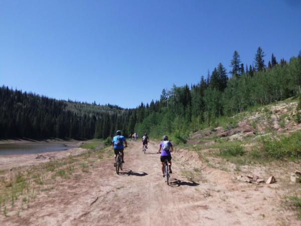 A group of cyclists riding along a dirt path near a lake, surrounded by lush greenery and tall trees under a clear blue sky. Turkey Flats mountain bike trail.