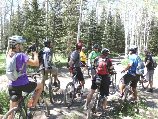 A group of mountain bikers in a forested area, gathered around a trail junction on a sunny day. They are wearing helmets and cycling gear, with some bicycles resting beside them. The background features tall trees and vibrant greenery, suggesting an outdoor adventure in nature. Turkey Flats mountain bike trail.