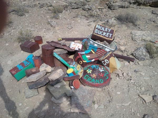A collection of colorful, painted objects arranged on a rocky surface, including a metal bucket labeled "Holey Bucket" and various other containers and stones, set in a dry, outdoor environment. Lunch Loops mountain bike trail.