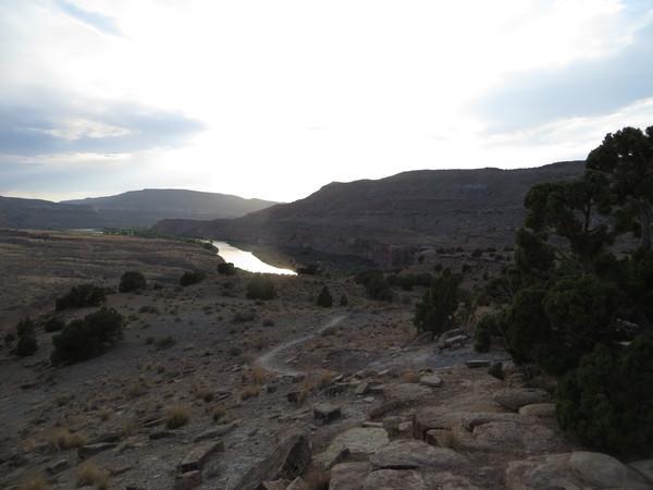 A scenic view of a river winding through rugged terrain, with green vegetation visible along the banks. The sun is setting behind distant hills, casting a soft light over the landscape. The foreground features rocky ground and sparse vegetation, creating a serene natural setting. Kokopelli Area Trails mountain bike trail.