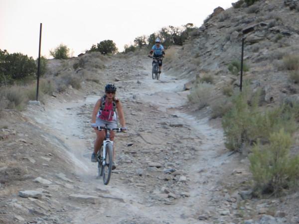 Two mountain bikers navigate a rocky trail surrounded by dry shrubs and vegetation. The first biker, wearing a helmet and a pink tank top, is riding downhill, while the second biker, dressed in a blue shirt, is further up the trail. The landscape shows a rugged terrain typical of outdoor biking locations. Kokopelli Area Trails mountain bike trail.