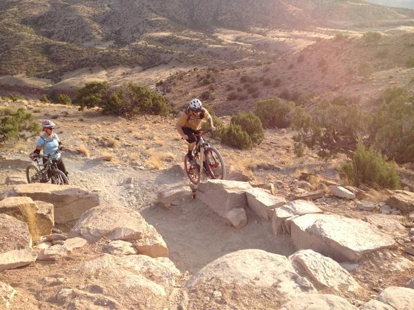 Two mountain bikers navigate a rocky trail in a mountainous landscape. One rider, wearing a helmet and a yellow shirt, is positioned on a rocky section, demonstrating skill while descending. The other rider, dressed in a blue shirt and helmet, is visible in the background, preparing to follow. The terrain features dry, sandy ground and sparse vegetation, with rolling hills in the distance under a bright sun. Kokopelli Area Trails mountain bike trail.